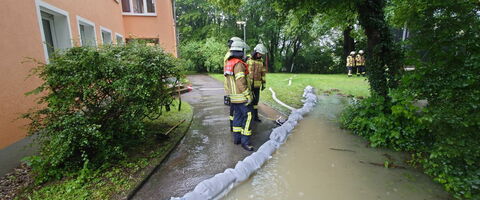 Feuerwehrleute sichern ein überflutetes Gebiet mit Sandsäcken nach einem Unwetter nahe eines Gebäudes.