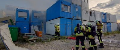 Feuerwehrleute bekämpfen einen Brand in einem Containergebäude, während Rauch in den Himmel aufsteigt.