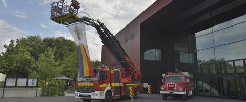Ein modernes Feuerwehrfahrzeug hebt eine Fahne vor einem Gebäude, daneben steht ein historischer Feuerwehrauto.