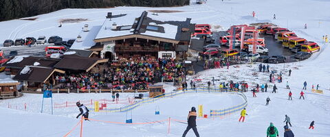 Skifahrer genießen einen sonnigen Tag auf der Piste neben einem belebten Bergrestaurant mit vielen Gästen.