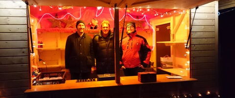 Drei Männer stehen in einem festlich beleuchteten Weihnachtsmarktstand und lächeln in die Kamera. | © FFW Unterföhring