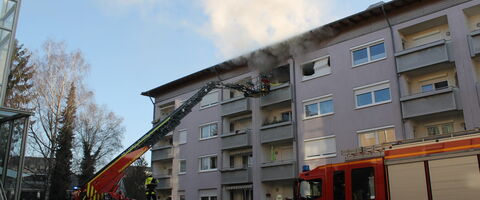 Feuerwehrleute bekämpfen einen ausgedehnten Zimmerbrand in einem Mehrfamilienhaus mit einem Leiterwagen.