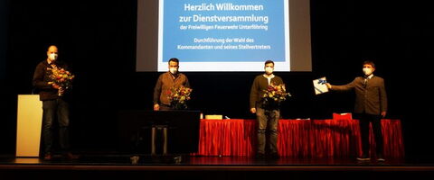 Bei der Kommandantenwahl 2021 stehen vier Männer mit Blumen und einem Dokument vor einer Leinwand auf einer Bühne.