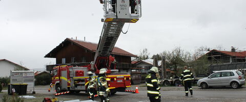 Feuerwehrleute arbeiten an einem Einsatzort mit einer Drehleiter vor einem Feuerwehrfahrzeug in einer ländlichen Gegend.